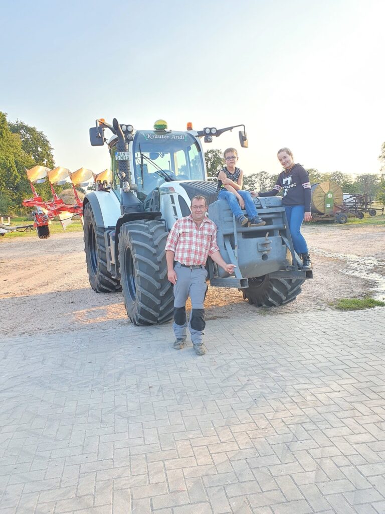 Ein Mann in kariertem Hemd und Arbeitshose steht vor seinem Traktor mit der Aufschrift Kräuter Andi, vorne auf dem Traktor sitzr ein Junge und steht ein Mädchen.